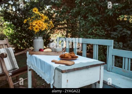 Table rustique sous les arbres dans le jardin. Nappe, fleurs jaunes et plateau avec de la nourriture dessus. Banque D'Images