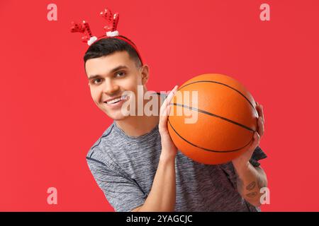Jeune homme heureux dans des cornes de renne avec basket-ball sur fond rouge Banque D'Images