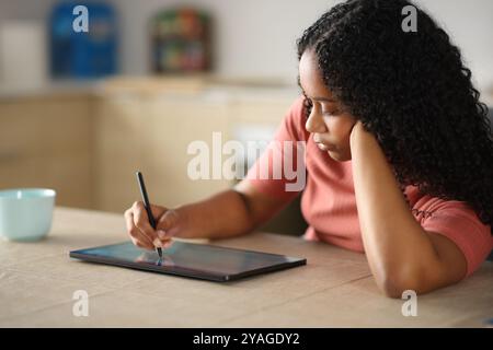 Femme sérieuse noire dessinant sur tablette numérique assise dans la cuisine Banque D'Images