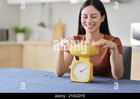 Femme asiatique heureuse pesant de la nourriture avec une balance dans la cuisine à la maison Banque D'Images