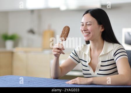 Heureuse femme asiatique tenant la crème glacée au chocolat et la regardant dans la cuisine Banque D'Images