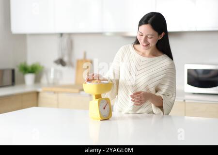 Femme asiatique heureuse pesant des céréales avec balance debout dans la cuisine à la maison Banque D'Images