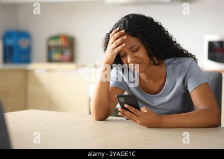 Femme triste vérifiant le téléphone assis dans la cuisine à la maison Banque D'Images