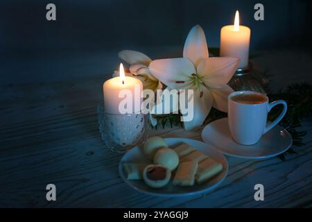 Deux bougies, fleurs de lis blanc, tasse à café et pralines, lumière festive sur fond bleu foncé, nature morte pour l'Avent, Noël et hiver Banque D'Images