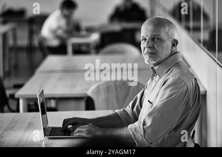 Image en noir et blanc d'un professionnel de l'informatique expérimenté travaillant sur son ordinateur portable dans un environnement de bureau occupé, mettant en valeur son expérience et sa concentration Banque D'Images