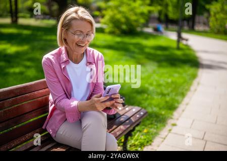Portrait de femme mature dans le parc. Elle envoie des messages sur son smartphone. Banque D'Images