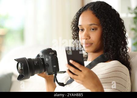 Photographe noir à la maison vérifiant téléphone et appareil photo reflex Banque D'Images