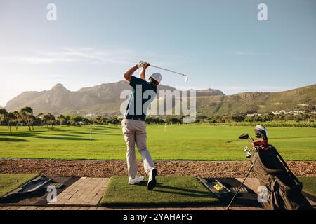 Golfeur masculin prenant un swing sur un beau parcours de montagne dans un complexe de golf, son sac de golf debout à proximité. Parfait pour les amateurs de golf et l'amour de la nature Banque D'Images