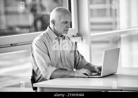 Homme d'affaires mature se concentre sur son travail, tapant sur un ordinateur portable dans un cadre de bureau lumineux et moderne. L'image en noir et blanc évoque une impression de pr Banque D'Images