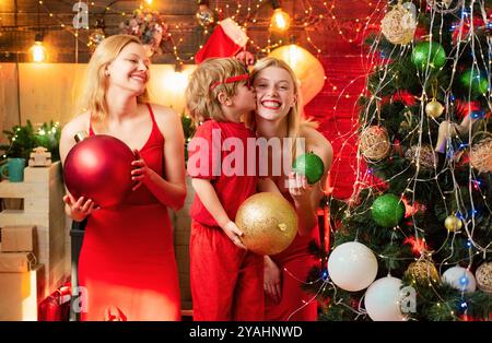 Joyeux Noël à la famille. Deux jeunes filles blondes en robe rouge jouent avec un petit garçon et ont beaucoup de temps. Souvenirs d'enfance. Vacances d'hiver en famille et Banque D'Images