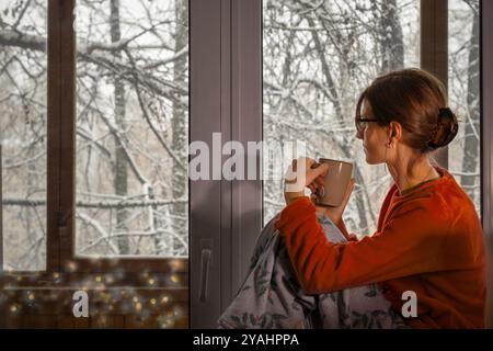 Jeune femme en pyjama maison en peluche confortable se trouve avec tasse par fenêtre et admire le paysage d'hiver avec des arbres enneigés. Noël Banque D'Images