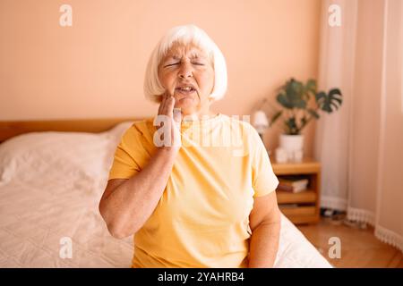 Malheureux caucasien triste des années 60 70 femme âgée avec mal de dents est assise sur un lit dans le salon. Femme retraitée en chemise jaune tenant la main sur son visage Banque D'Images