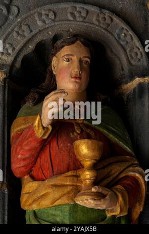 Saint Jean l'Apôtre tient une coupe d'or. Tête et corps supérieur d’une des 12 statues d’apôtres polychromes, sculptées en bois en 1721, qui flanquent l’entrée d’une église bretonne, L’Eglise Saint Sauveur au Faou, Finistère, Bretagne, France. Les statues ont été récemment restaurées et repeintes. Banque D'Images