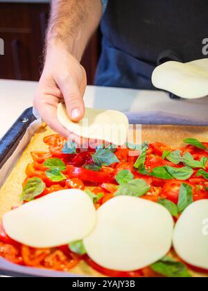 jeune homme mettant du fromage blanc sur une pizza avec des tomates fraîches et des feuilles de basilic Banque D'Images