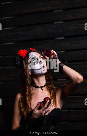 La jeune femme pose avec un crâne de sucre, une robe noire et des roses rouges devant un fond de bois foncé. Elle célèbre le jour des morts Banque D'Images