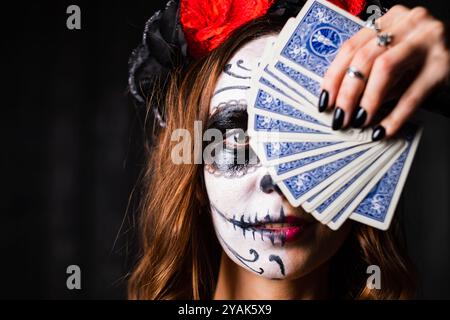 Femme couvre une partie de son visage avec des cartes à jouer, montrant son maquillage de crâne de sucre dia de los muertos. Elle a les cheveux rouge foncé et porte un voile noir et une couronne de fleur rouge Banque D'Images
