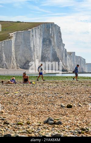 Les sept Seven Sisters avec leurs falaises de craie se dressent en grand tandis que deux hommes passent devant des femmes bronzées sur la plage de Cuckmere Haven dans le Sussex de l'est, en Angleterre. Banque D'Images