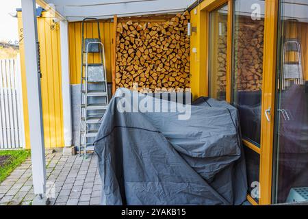 Bois soigneusement empilé sous le patio couvert à côté de la villa jaune, avec échelle et mobilier de jardin recouvert de bâche, préparé pour la saison d'hiver. Suède. Banque D'Images