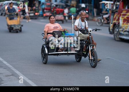 12/10/2024 Cebu City, Philippines. Un homme conduisant un tricycle électrique moderne. Une femme est assise du côté passager avec ses courses. Ces ec Banque D'Images