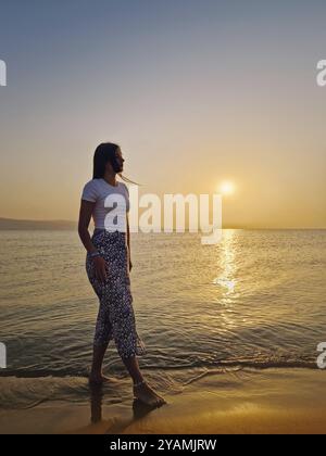 Jeune femme esthétique marchant pieds nus dans l'eau sur la plage à l'aube, regardant le lever de soleil chaud au-dessus de la mer. Belle scène matinale bord de mer, su Banque D'Images