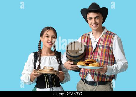 Jeune couple en vêtements traditionnels allemands avec tonneau de bière, saucisses et bretzels sur fond bleu. Concept Octoberfest Banque D'Images