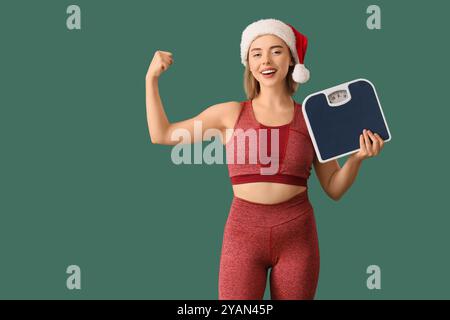 Jeune femme sportive en chapeau de Père Noël avec des écailles montrant des muscles sur fond vert Banque D'Images