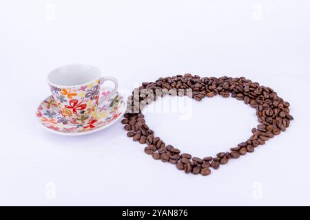 Une forme de coeur formée par des grains de café à côté d'une tasse de café à la vapeur, sur un fond blanc propre Banque D'Images