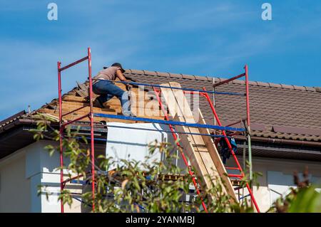 Ouvrier réparant des tuiles sur une maison, debout sur un échafaudage sous un ciel bleu clair. Banque D'Images
