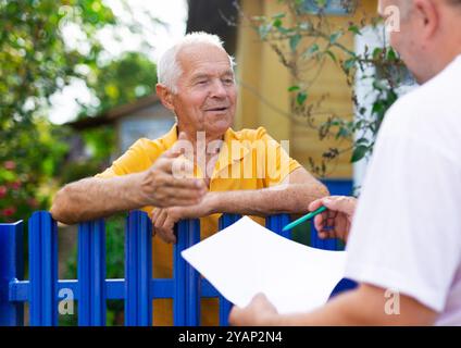 Homme senior communiquant avec le représentant de la compagnie d'assurance tout en se tenant à la clôture de sa maison de campagne Banque D'Images