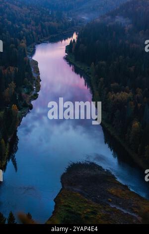 Une rivière sinueuse coule à travers une vallée boisée luxuriante, reflétant les douces teintes crépusculaires dans ses eaux calmes alors que le soleil se couche au loin, créant un pois Banque D'Images