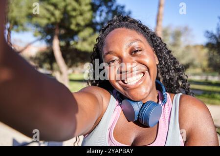 Femme afro-américaine joyeuse prenant selfie sur la pause de l'entraînement, portant des écouteurs et souriant à la caméra. Heureuse dame faisant photo d'elle-même sur tra Banque D'Images