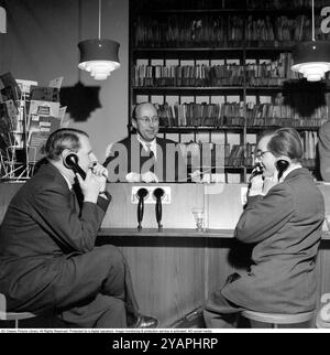 Comment vous avez acheté des disques dans les années 1950 Les hommes tenant ce qui ressemble à un téléphone écoutent en fait des enregistrements. À cette époque, il était courant que les clients puissent écouter les disques dans le magasin avant de décider de les acheter. Le magasin de disques dispose de plusieurs stations d'écoute et le personnel derrière le comptoir gère le système. Ce magasin particulier est Selanders skivbar sur Vasagatan à Stockholm. 1955. Banque D'Images