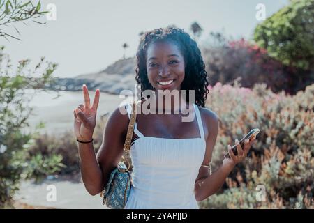 Une femme sourit et tient un téléphone portable tout en se tenant debout dans un champ. Elle porte une robe blanche et un sac à main Banque D'Images