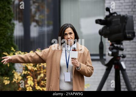 Une journaliste multiraciale vêtue d'un manteau beige se tient confiante, tenant un micro et diffusant en direct. Banque D'Images