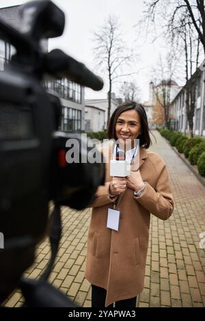 Une journaliste multiraciale se tient en toute confiance dehors, tenant un micro, prête à rapporter des nouvelles. Banque D'Images