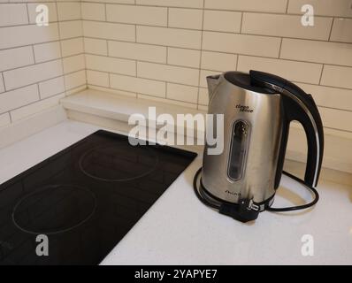Une bouilloire à thé argentée se trouve sur un comptoir blanc à côté d'un poêle noir. La bouilloire est branchée sur le poêle et le poêle est éteint. La scène est sim Banque D'Images