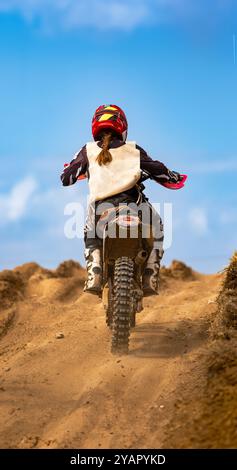 Vue arrière d'une pilote de motocross femme en action sur le sommet d'une colline sur une piste de terre lors d'une course d'endurance de motocross, dégageant la poussière et le gravier esprit Banque D'Images