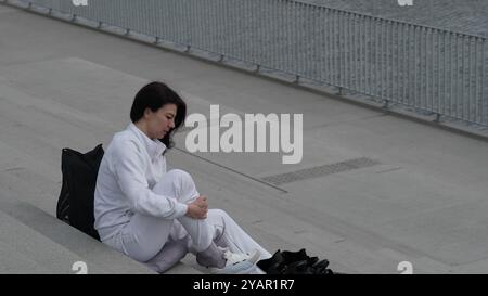 Une femme en vêtements athlétiques blancs s'assoit sur des marches ajustant ses chaussures de saut. La scène met l'accent sur la préparation et le dévouement à la forme physique en milieu urbain Banque D'Images