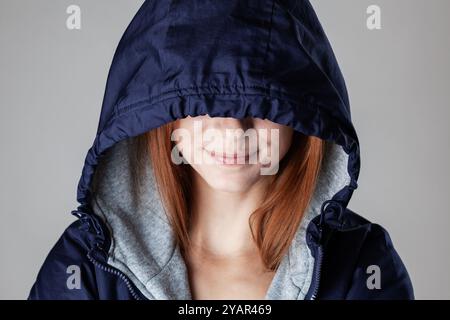 Portrait en gros plan d’une jeune femme aux cheveux roux, partiellement dissimulée par sa capuche, présentant un sourire subtil. Idéal pour le style de vie et la mode. Banque D'Images