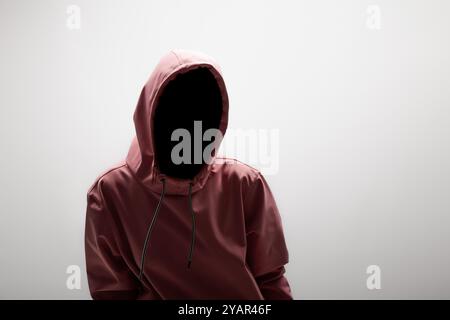 Mystérieux et moderne : une jeune femme dans un sweat à capuche rose, visage obscurci, posant sur un fond gris doux. Banque D'Images