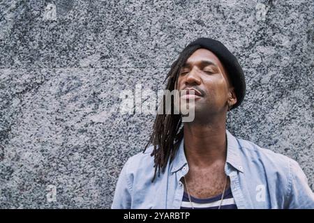 Bel homme afro-américain en tresses avec béret élégant regardant loin tout en se tenant debout contre le mur gris et les yeux fermés Banque D'Images