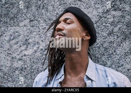 Bel homme afro-américain en tresses avec béret élégant regardant loin tout en se tenant debout contre le mur gris et les yeux fermés Banque D'Images