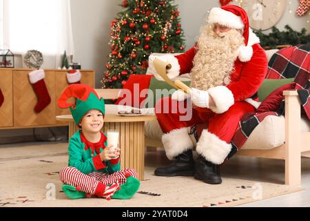 Père Noël lisant lettre et mignon petit elfe buvant du lait à la maison la veille de Noël Banque D'Images