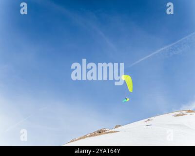 Un parapente avec une aile orange éclatante survole des montagnes enneigées à serre Chevalier, Briançon, France. Le ciel lumineux et les pics accidentés créent Banque D'Images