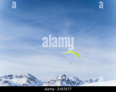 Un parapente avec une aile orange éclatante survole des montagnes enneigées à serre Chevalier, Briançon, France. Le ciel lumineux et les pics accidentés créent Banque D'Images