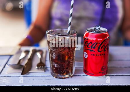 Verre de cola avec de la glace et de la paille rayée à côté d'une boîte de Coca-Cola sans sucre sur la table avec des ustensiles. Banque D'Images