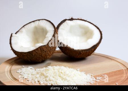 Portion de noix de coco râpée déshydratée sur une planche de bois ronde et deux moitiés de noix de coco coupées en deux avec la pulpe dans le fond et hors de Banque D'Images