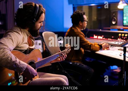 Musicien composant de nouvelles pistes sur sa guitare électro-acoustique, jouant disque sur instrument pour créer un album. Chanteur collaborant avec le producteur pour faire de nouveaux disques en salle de contrôle au studio. Banque D'Images