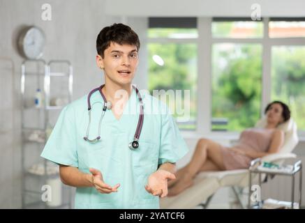 Jeune homme médecin posant dans le cabinet du médecin avec le patient allongé sur le fauteuil clinique Banque D'Images