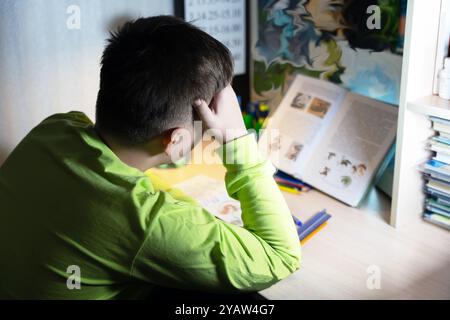 Un garçon fait ses devoirs à son bureau. Il pose la tête sur sa main et lit un manuel. La table est éclairée par une lampe. Banque D'Images
